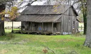 An abandoned single-story home in rural Tunica County, Mississippi, with a sagging roofline and weathered wood siding against an overcast sky. Contemporary photograph of housing stock in the county that ranks second of 3,144 nationally for household financial distress.